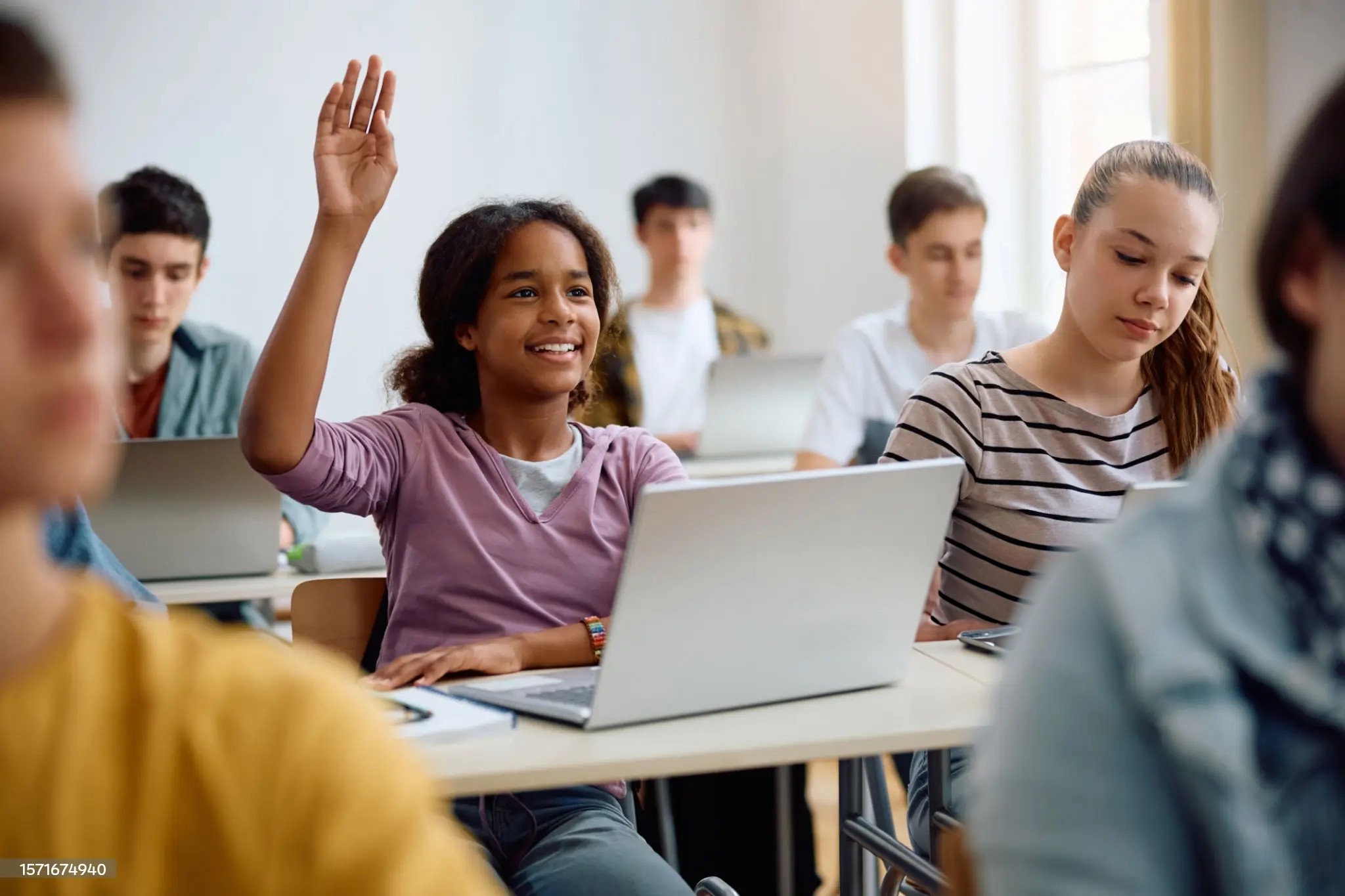 Happy African American teenage girl using laptop and raising her hand to ask a question during computer class at high school.